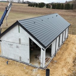 Ostafiński Mariusz Usługi Dekarsko - Ciesielsko - Blacharskie - Montaż dachu z szarej dachówki na surowym budynku z użyciem dźwigu. Widok z góry na budynek w trakcie budowy, otoczony polem i lasem.