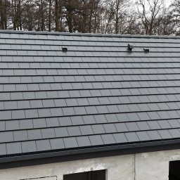 Ostafiński Mariusz Usługi Dekarsko - Ciesielsko - Blacharskie - Szary dach z dachówki ceramicznej, widoczne kominki wentylacyjne. Budynek w stanie surowym zamkniętym, drzewa w tle. Ujęcie z góry, lekko z boku.