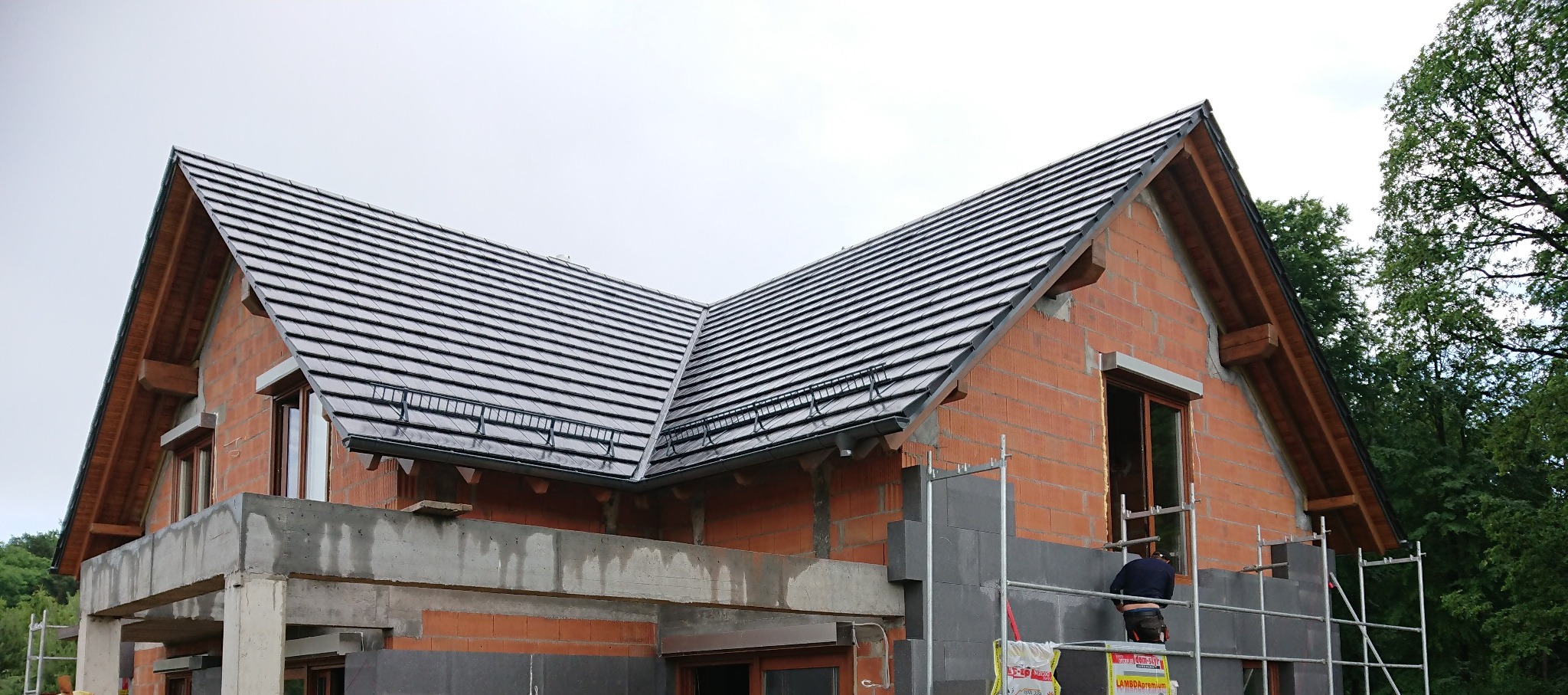 Budowa domu jednorodzinnego: czarna dachówka ułożona na częściowo wymurowanej konstrukcji, widoczne rusztowania i pracownik w trakcie wykonywania prac murarskich.