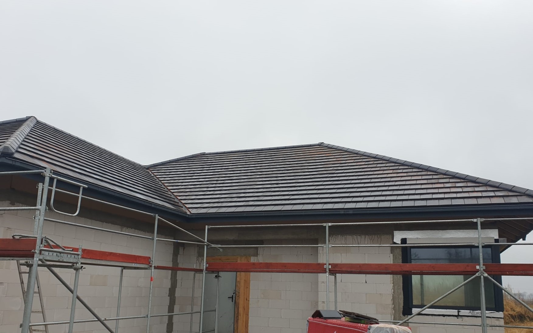 Nowy dach z ciemnej dachówki ceramicznej na budynku w trakcie budowy, widoczne rusztowania i niezakończone prace elewacyjne.