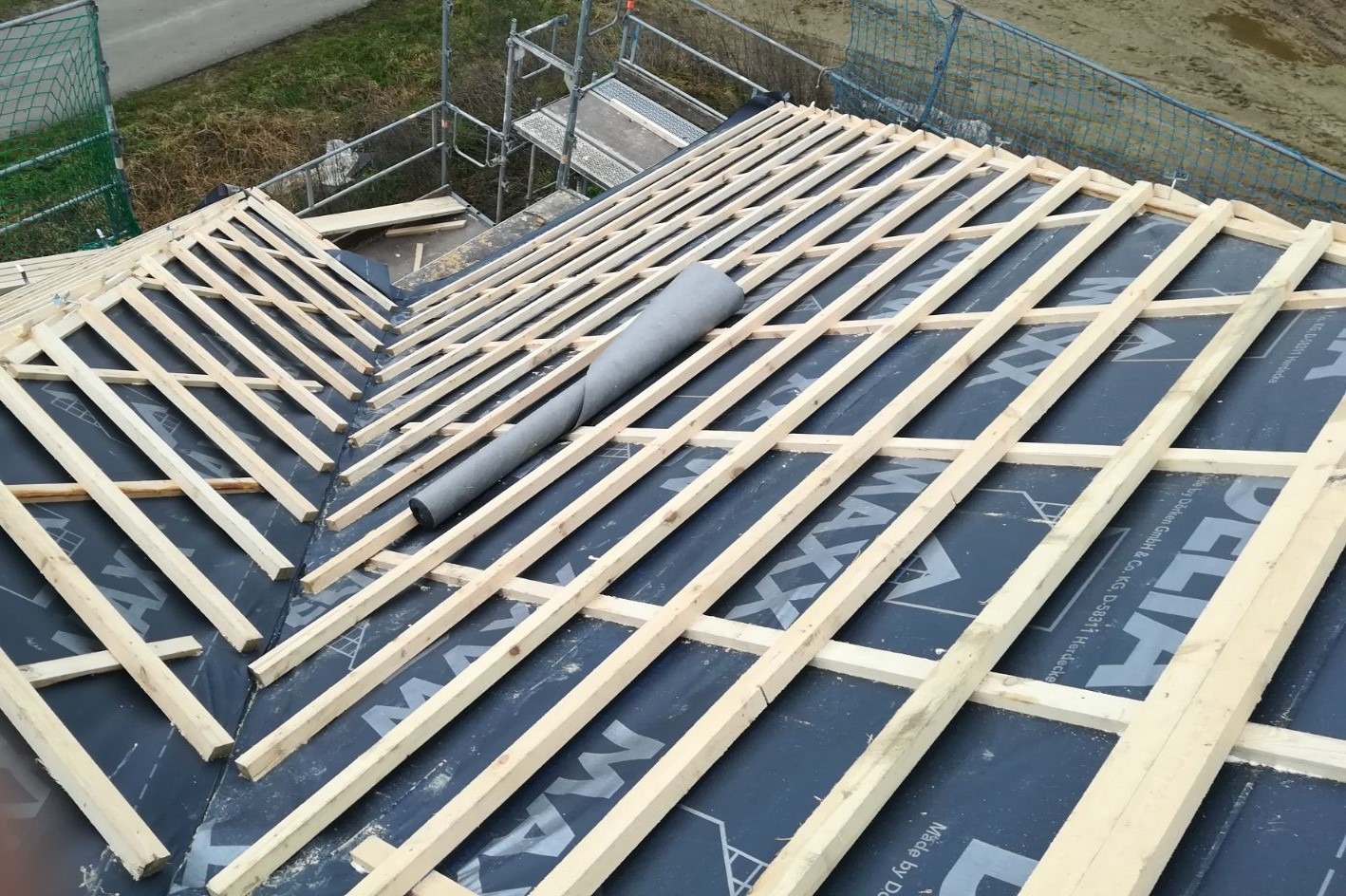 W trakcie budowy: Widok z góry na drewnianą konstrukcję dachu pokrytą czarną membraną dachową z widocznym fragmentem rolki izolacyjnej i rusztowaniem w tle.