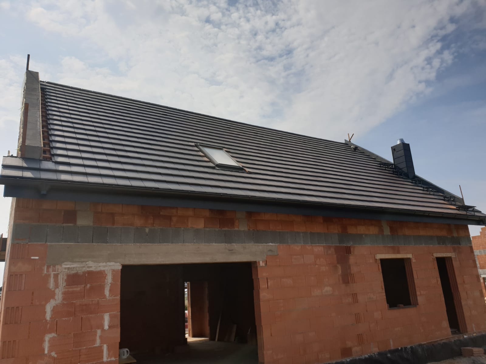 Nowoczesny dom w trakcie budowy, z ciemną dachówką ceramiczną, oknem dachowym i kominem. Mur z czerwonej cegły, widoczne nadproża okienne i garażowe.