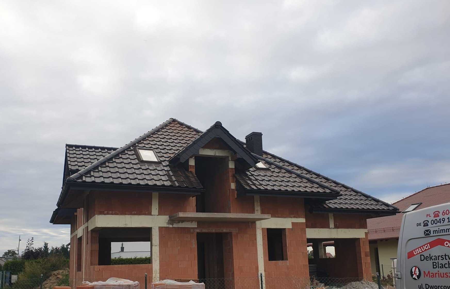 Dom w trakcie budowy z częściowo ukończonym, brązowym dachem z dachówki ceramicznej, widoczne okna dachowe i komin, ściany z czerwonej cegły w stanie surowym.