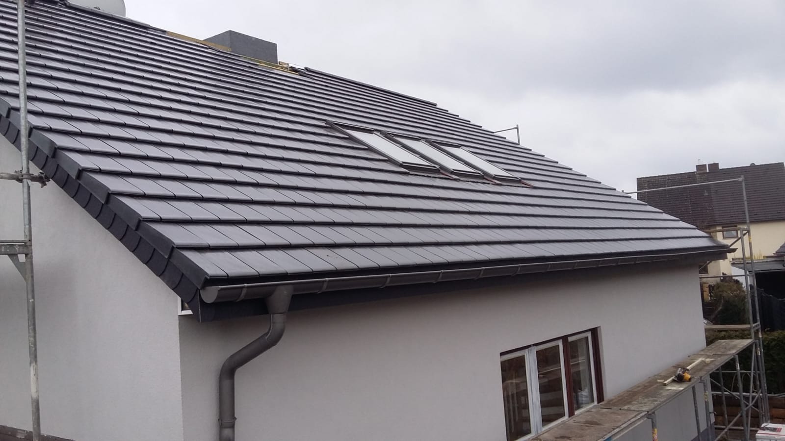 Nowy dach z ciemnoszarej dachówki ceramicznej z trzema oknami dachowymi, świeżo pomalowana elewacja budynku w jasnym odcieniu, zamontowane rynny, widoczne rusztowanie.