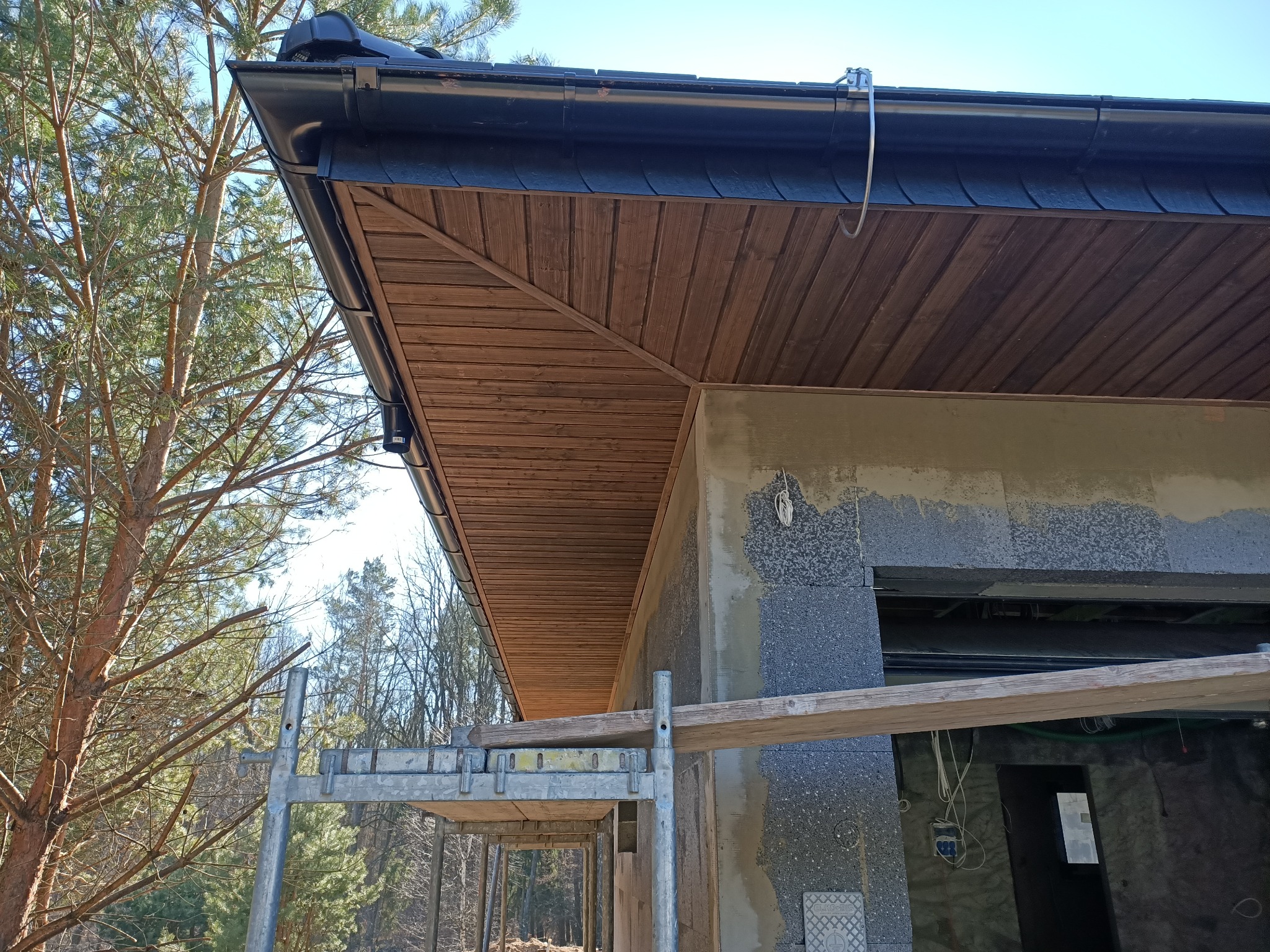 Drewniane podbitki dachowe z czarnymi rynnami i fragmentem rusztowania na tle drzew i błękitnego nieba.