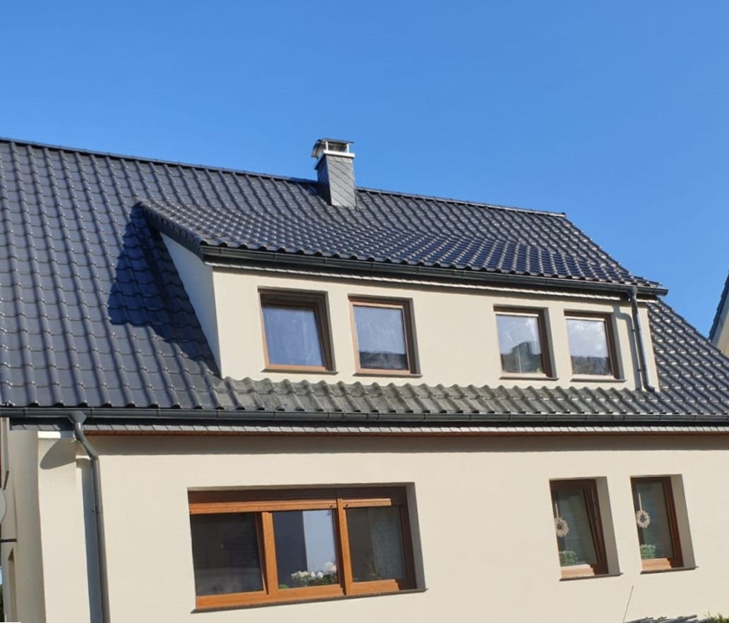 Szary dach z dachówki ceramicznej na jasnym domu jednorodzinnym, widoczne okna dachowe i komin z blachy, rynny w kolorze dachu.