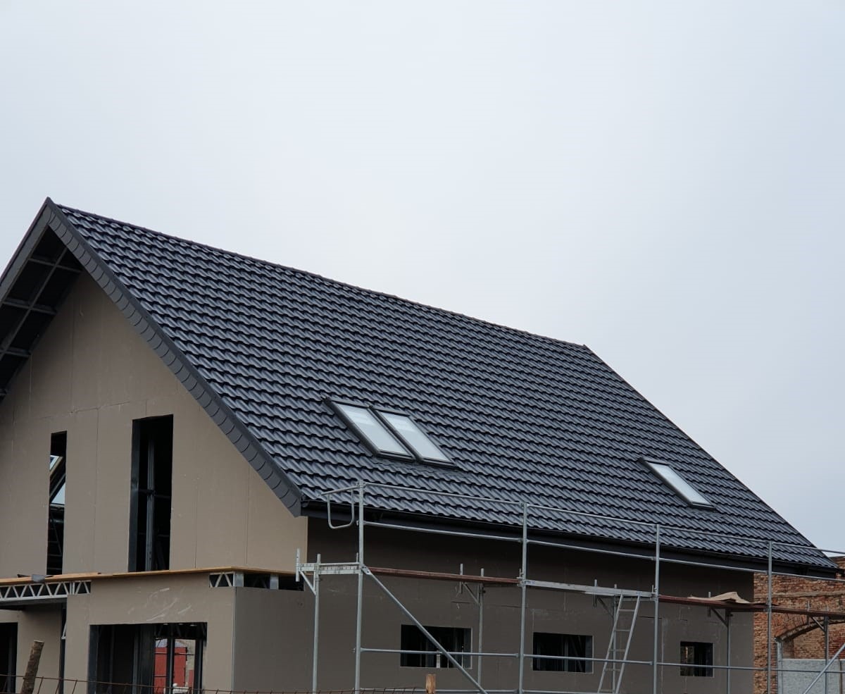 Dom w trakcie budowy z nowym, ciemnym dachem z dachówką i oknami połaciowymi, widoczne rusztowania.