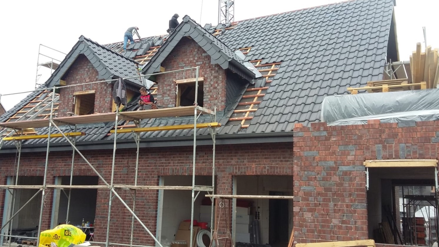 Montaż dachówki na nowo budowanym domu z cegły, widoczni dekarze pracujący na dachu i rusztowaniach, częściowo pokryty dach w ciemnoszarym kolorze.