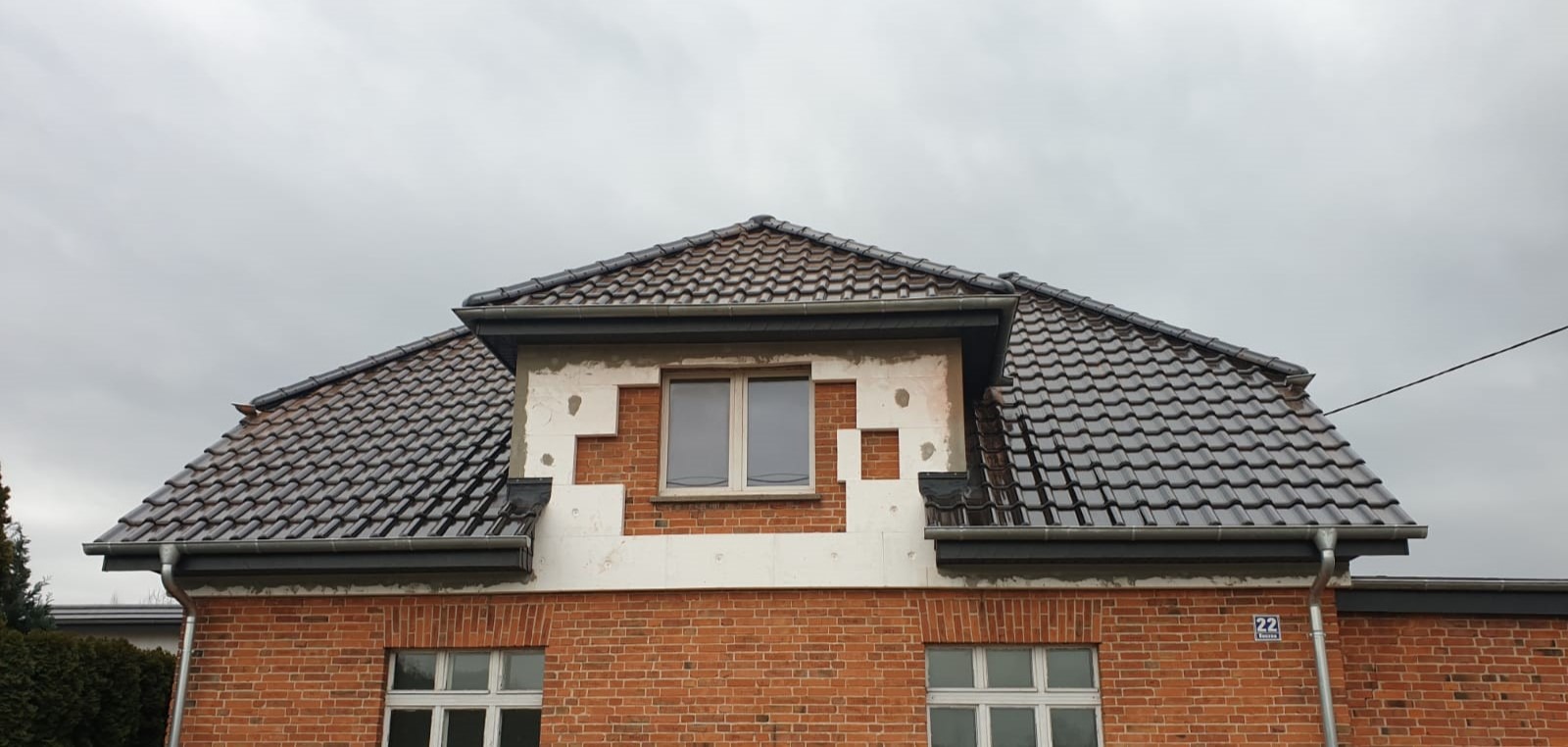 Dwuspadowy dach z ciemnej dachówki ceramicznej, widoczne okno dachowe z elementami elewacji w trakcie wykończenia, rynny z blachy w kolorze grafitowym.