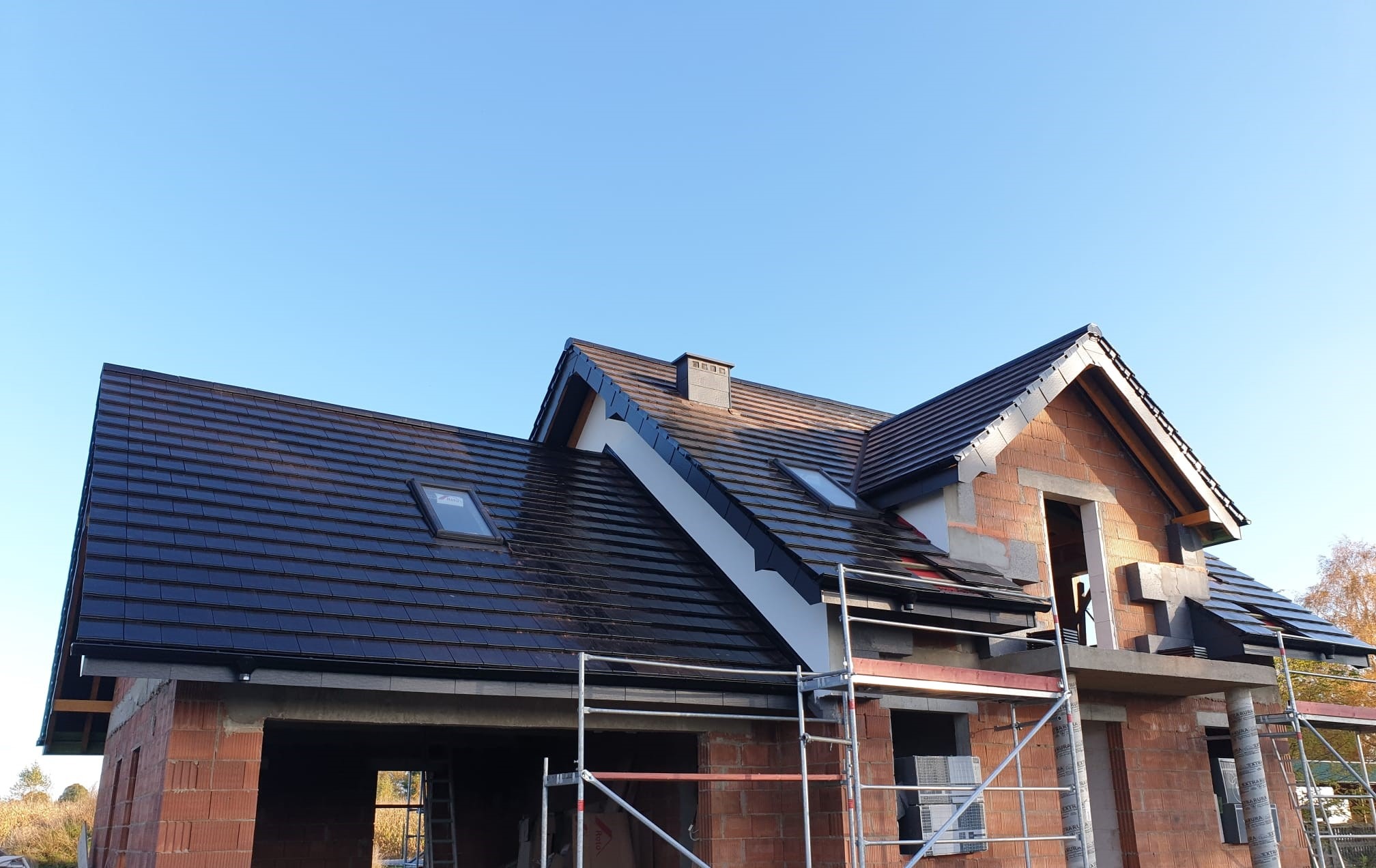 Dom w trakcie budowy z nowym, błyszczącym, ciemnym dachem z dachówkami ceramicznymi, widoczne okna dachowe i rusztowania.
