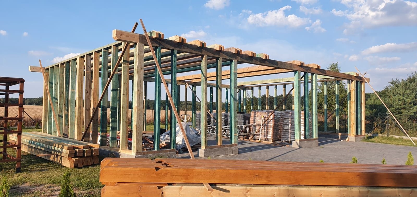 Konstrukcja drewniana szkieletowa w trakcie budowy, z elementami impregnowanymi na zielono, na tle błękitnego nieba z chmurami i pola pszenicy w oddali.