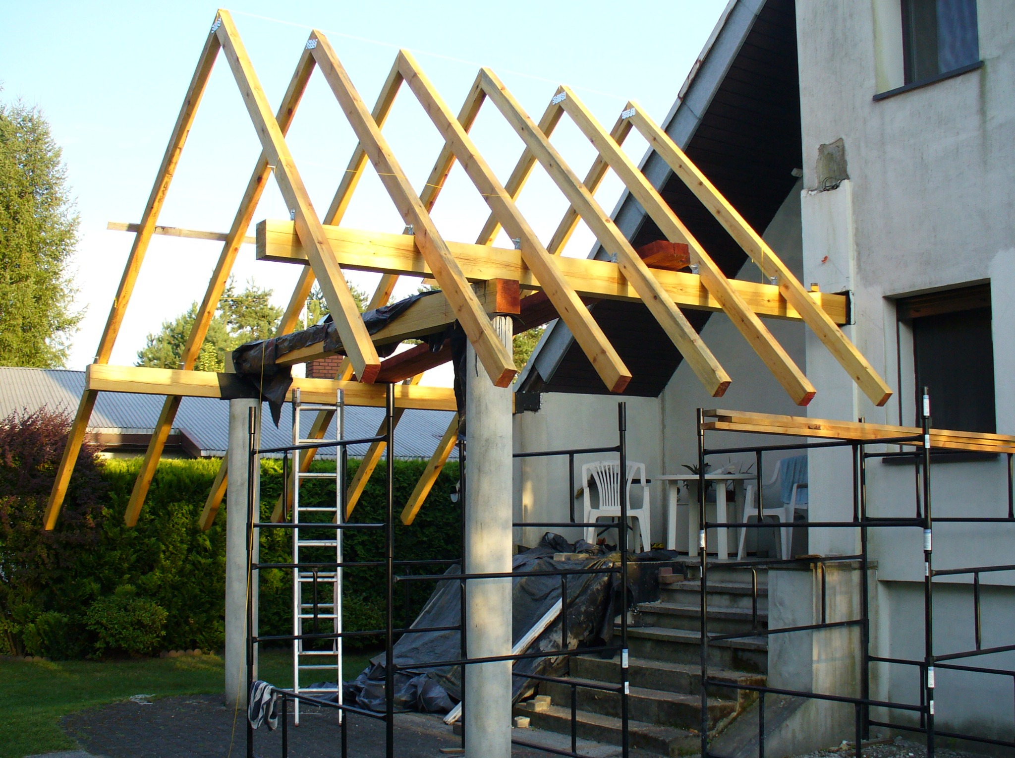 Wznoszenie drewnianej konstrukcji dachu na betonowym filarze, widoczne rusztowanie, drabina i fragment elewacji budynku w tle.