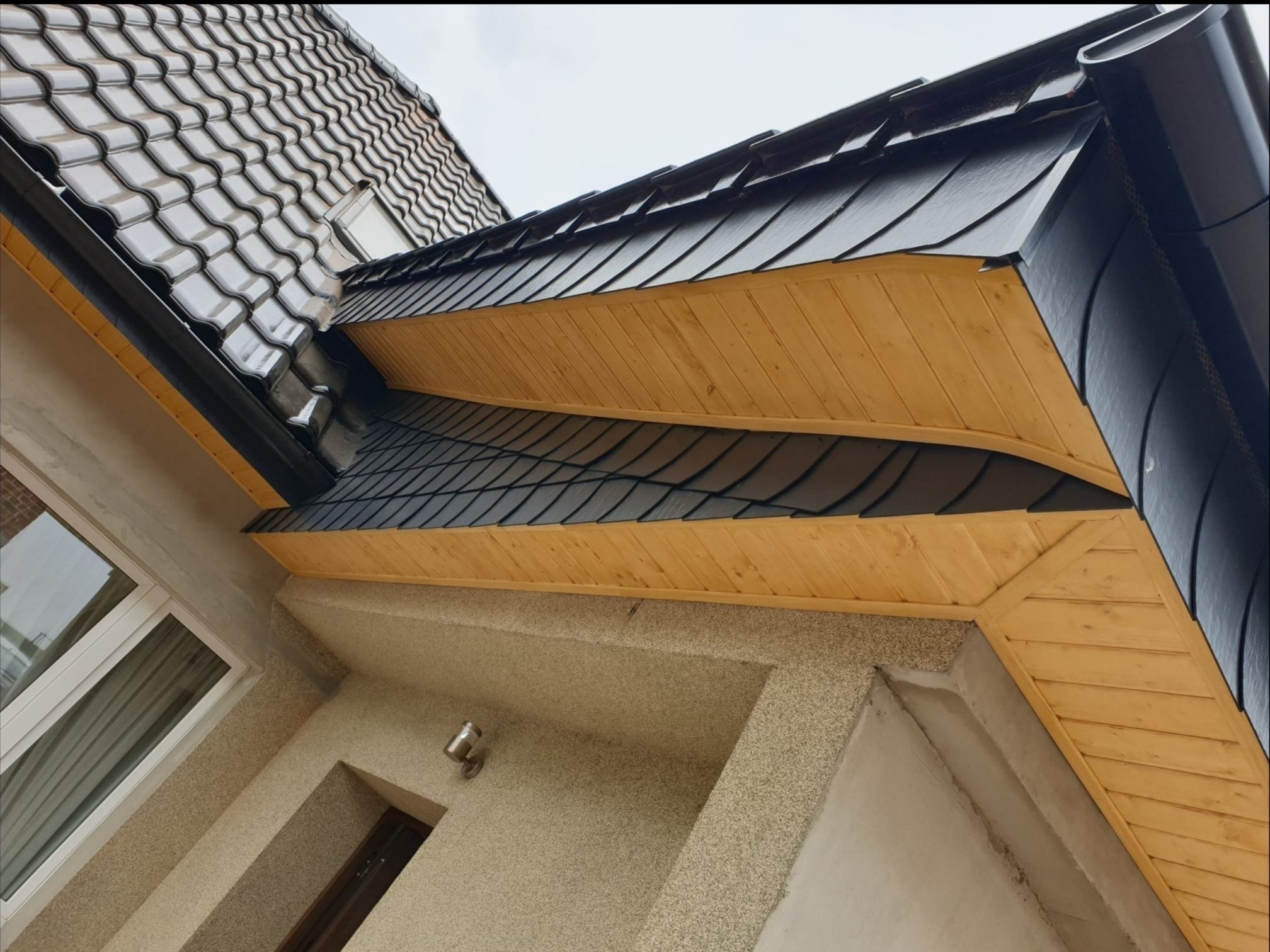 Nowoczesny dach z czarnej dachówki karpiówki, wykończony drewnianą podbitką w kolorze jasnego drewna, widok z dołu na elewację budynku.