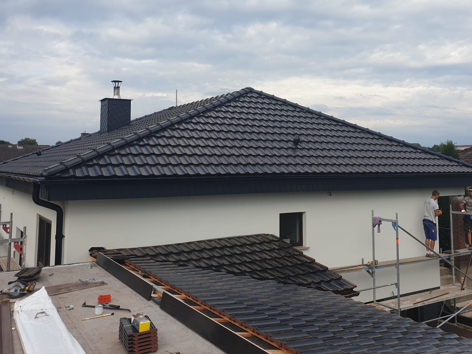Dom z częściowo zdjętym starym i nowym dachem z czarnej dachówki, ekipa montuje rynny na rusztowaniu.