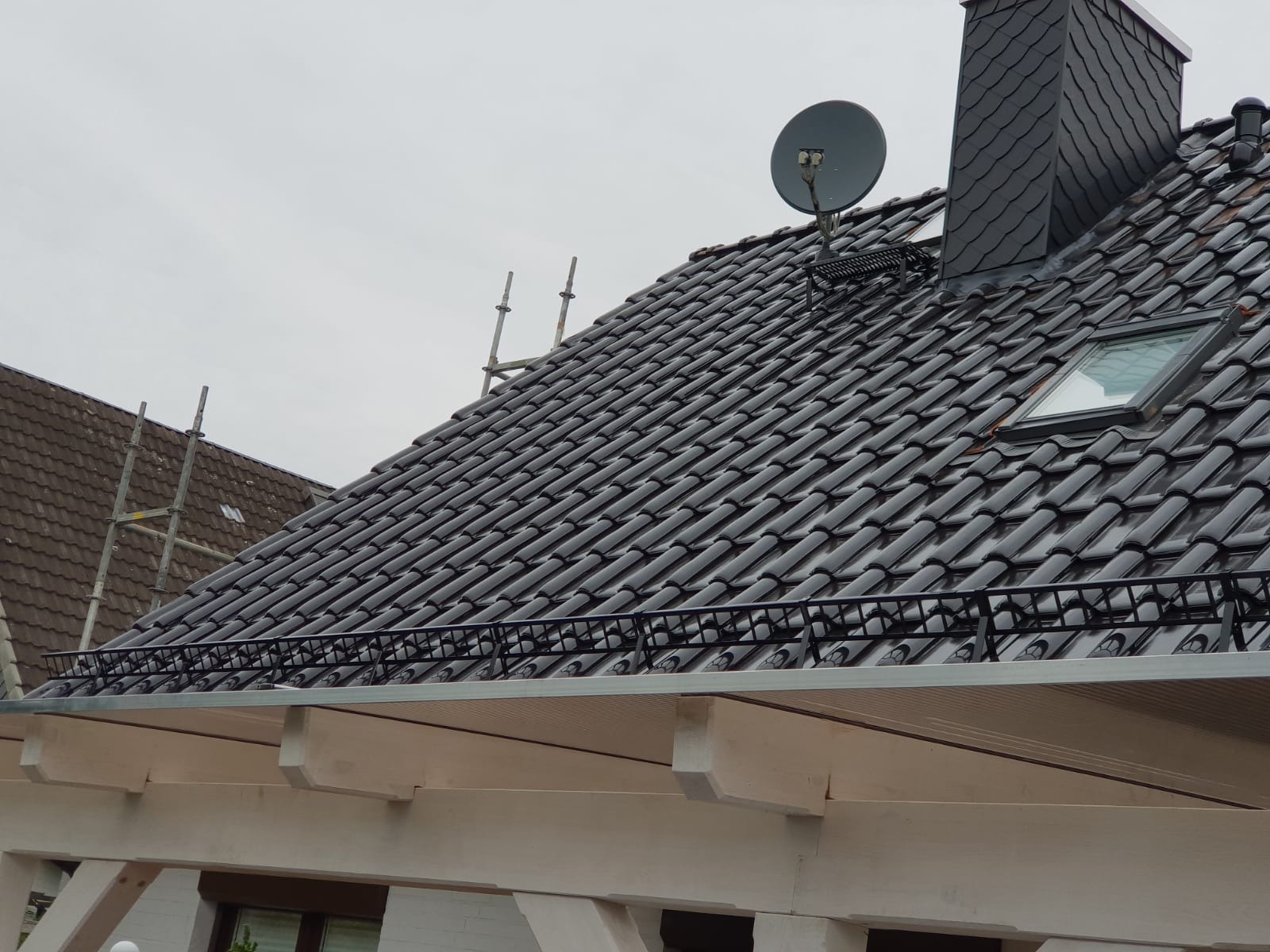 Błyszczący, czarny dach z dachówki ceramicznej z oknem dachowym i kominem z łupka, z widoczną anteną satelitarną, na tle pochmurnego nieba.