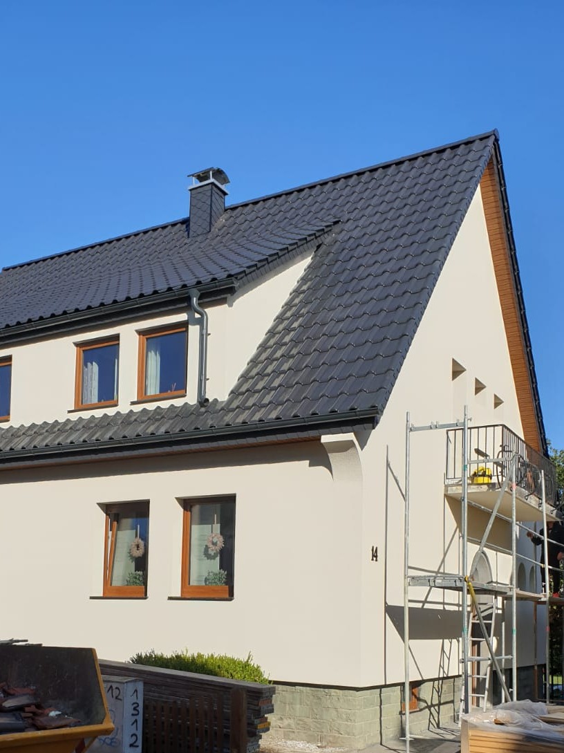 Dom jednorodzinny po remoncie dachu z nową, ciemną dachówką ceramiczną i rynnami, widoczne rusztowanie i kontener na odpady budowlane.