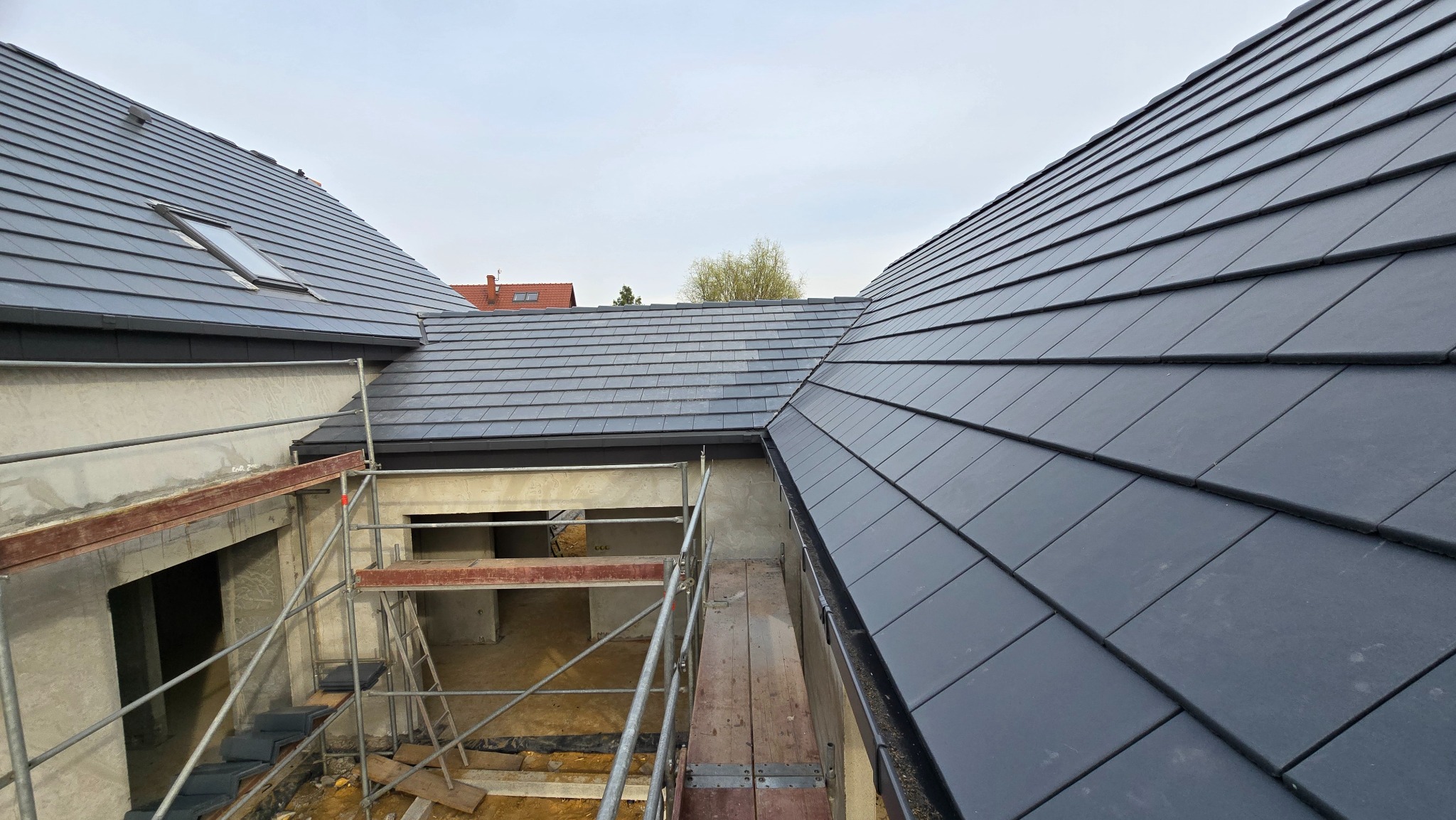 Nowy dach z szarej dachówki ceramicznej, widok z góry na budynek w trakcie budowy. Na pierwszym planie rusztowanie i fragment niezadaszonej konstrukcji.