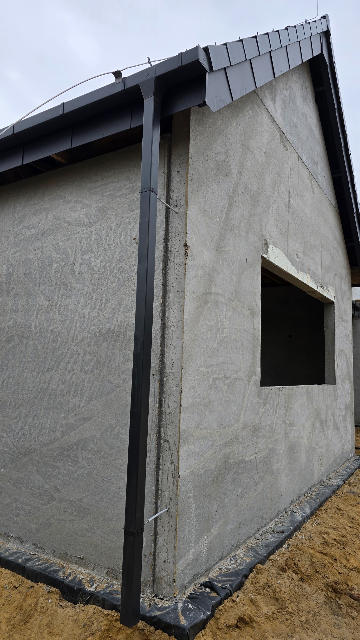 Surowy budynek w trakcie budowy z zamontowanymi rynnami w kolorze grafitowym i fragmentem dachu pokrytego dachówką. Widoczne okno bez ramy.