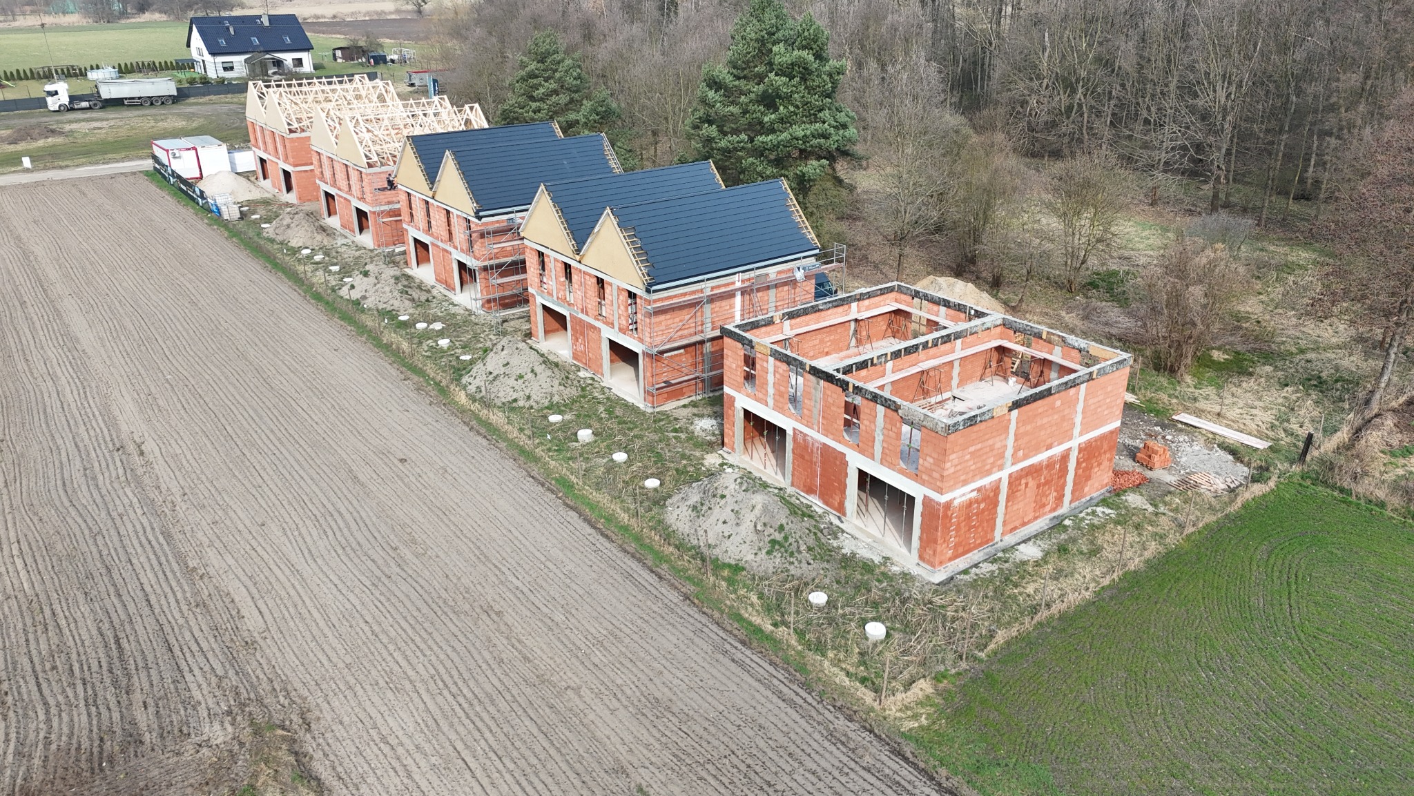 Budowa szeregu domów jednorodzinnych z czerwonej cegły, widok z lotu ptaka. Domy w różnym stadium zaawansowania: od fundamentów po konstrukcję dachu z pokryciem.