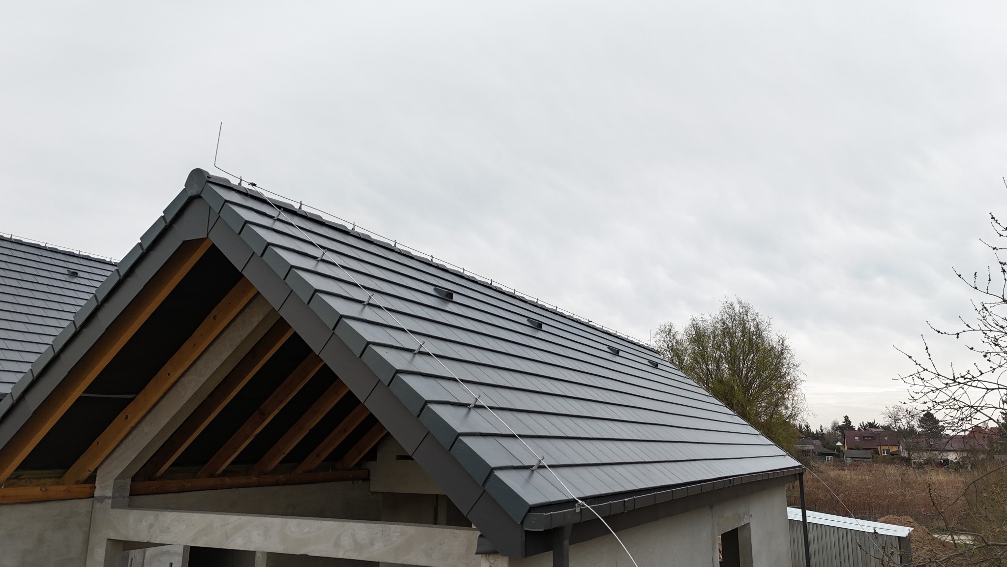 Szary dach z dachówki modułowej na surowym budynku w trakcie budowy, widoczna więźba dachowa i system odgromowy. Pochmurne niebo w tle.