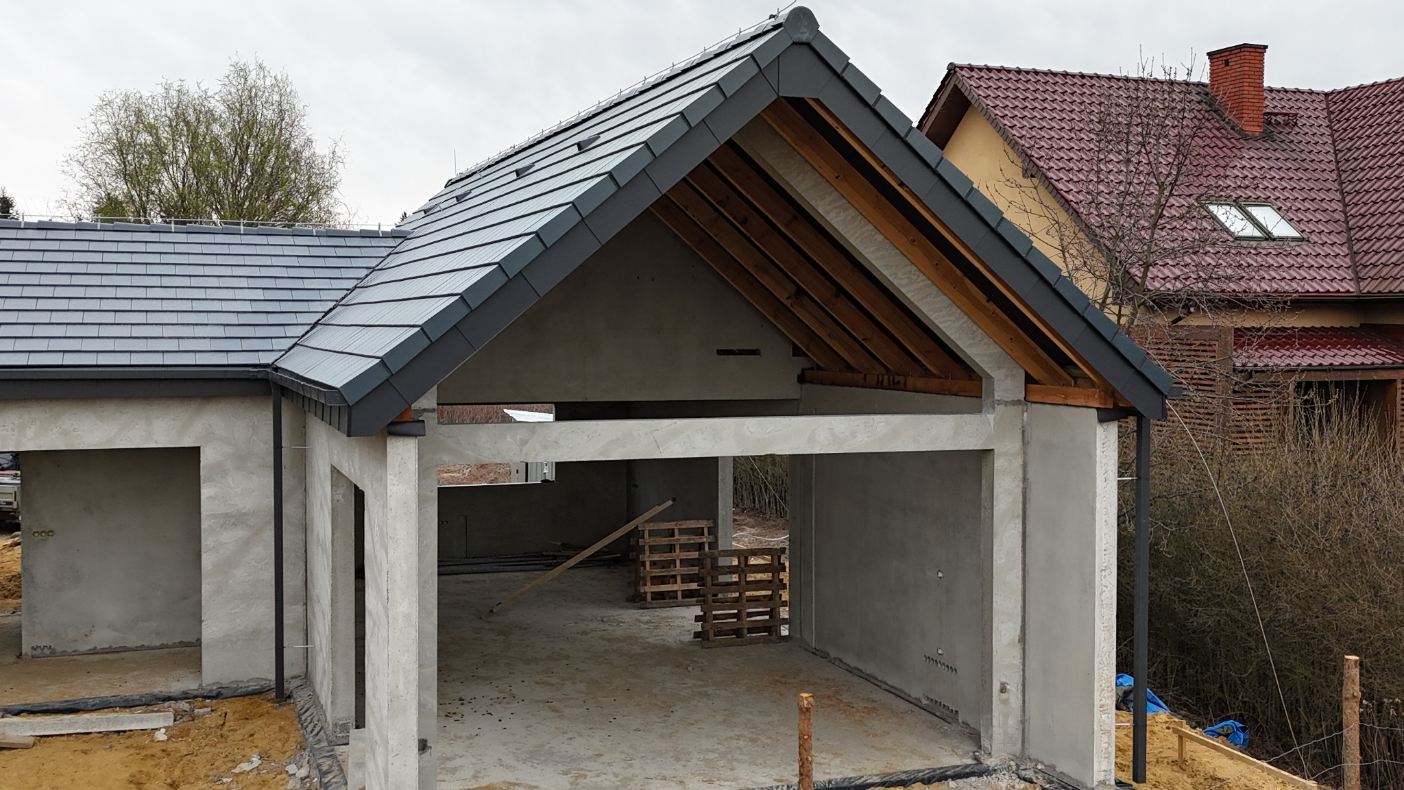 Surowy garaż z szarą dachówką karpiówką w trakcie budowy. Widoczna więźba dachowa i fragment dachu sąsiedniego budynku z czerwoną dachówką.