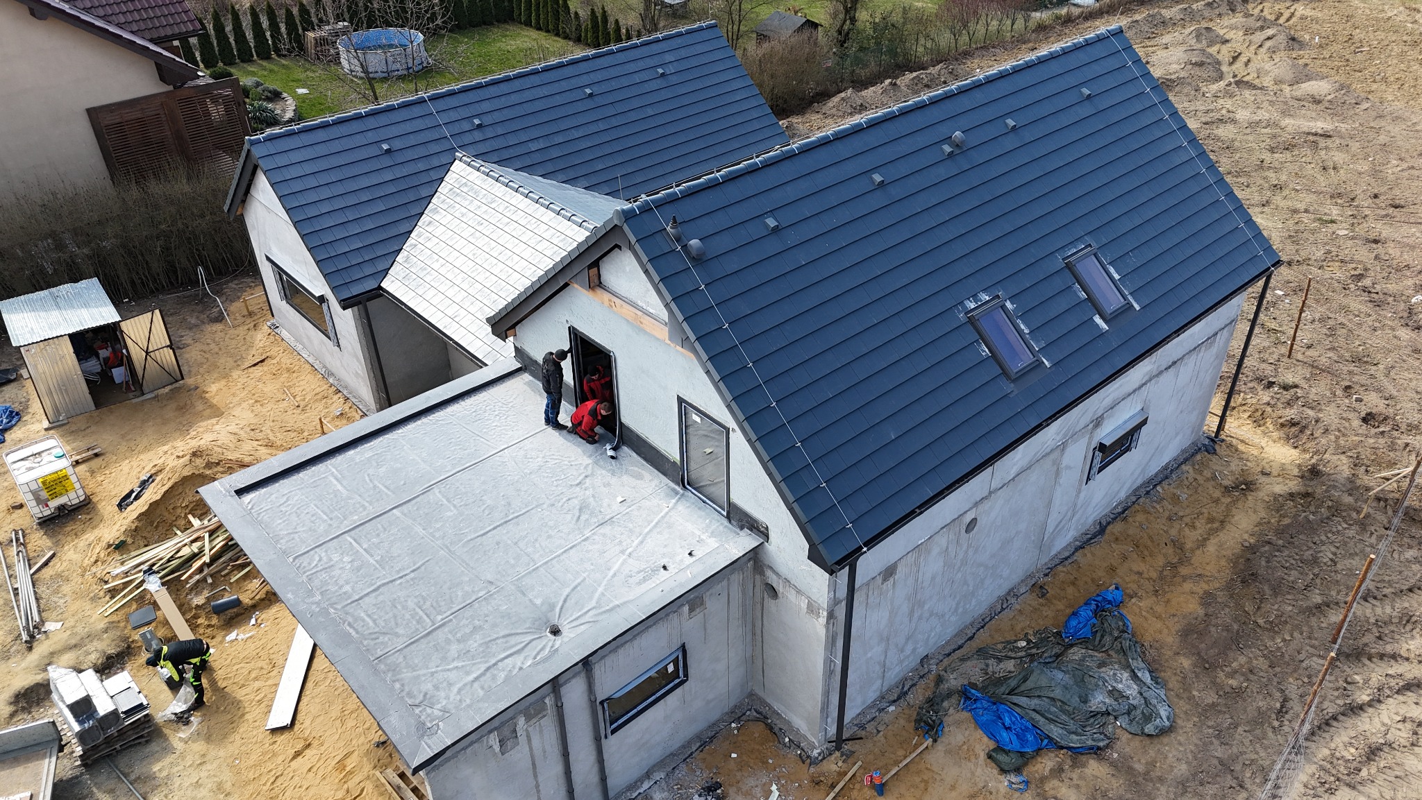 Nowoczesny dom w trakcie budowy z ciemną dachówką i oknami dachowymi. Widoczni pracownicy montujący elementy. Ujęcie z góry ukazuje postęp prac.