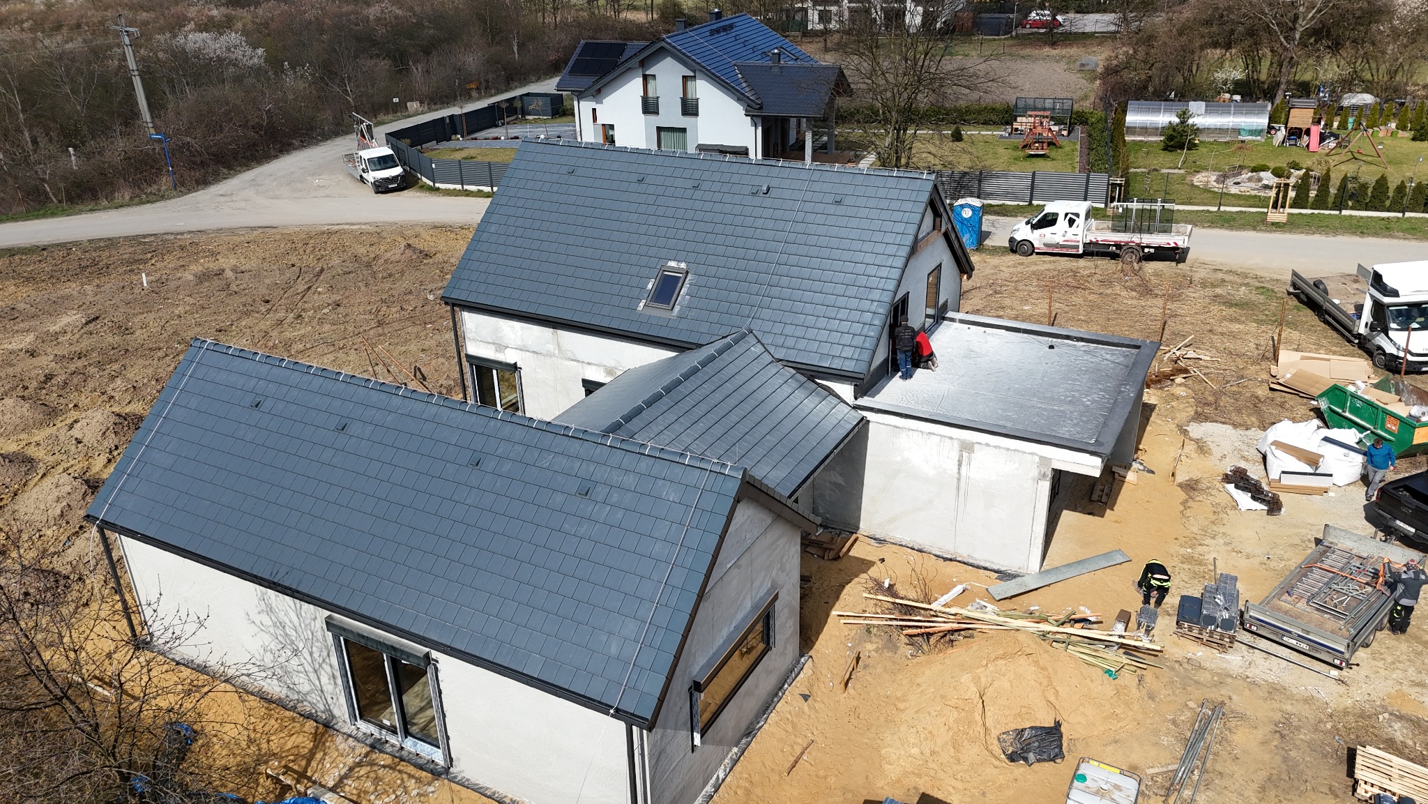 Widok z góry na dom w trakcie budowy z szarym dachem z dachówki. Na dachu pracują dwie osoby. W tle inne budynki i drzewa. Widoczne materiały budowlane.