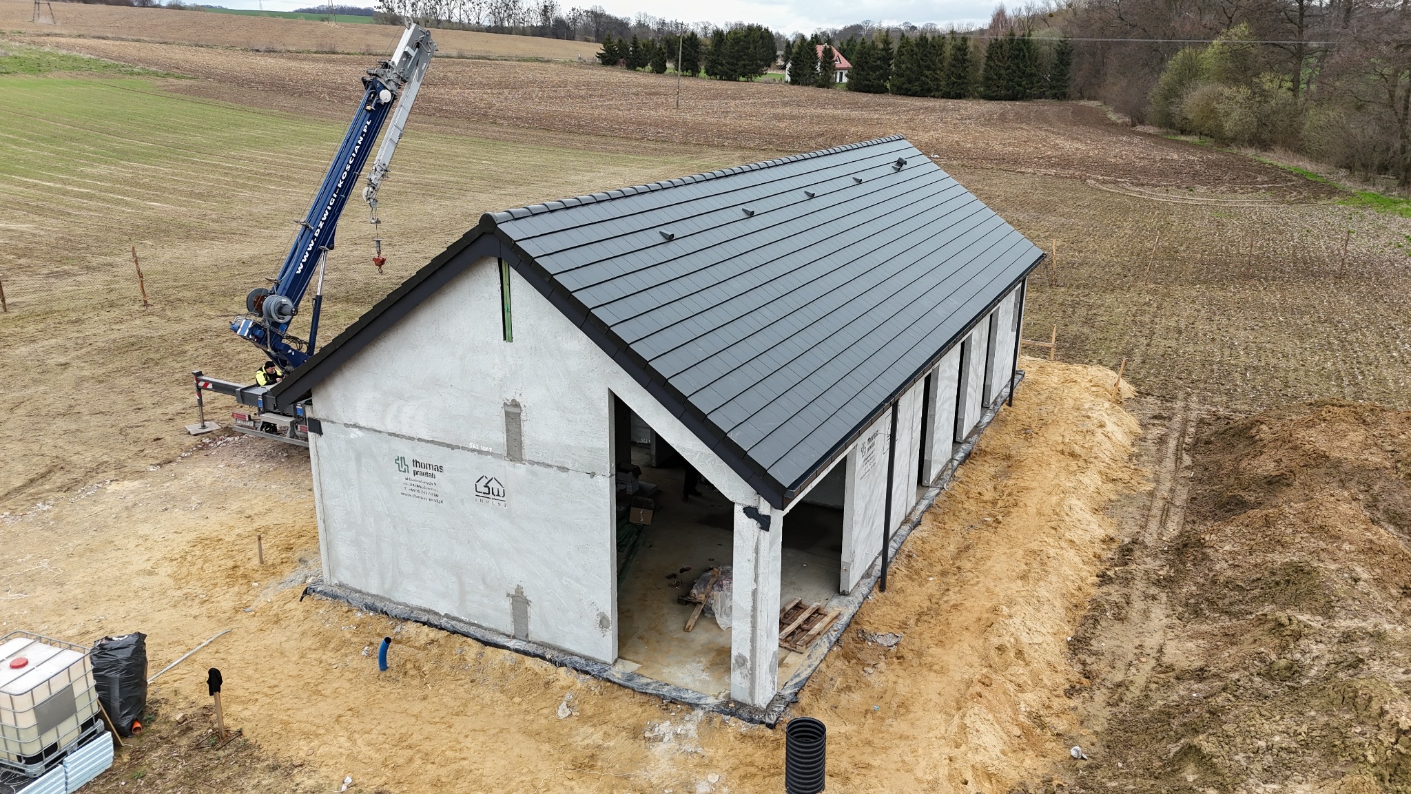 Montaż dachu z szarej dachówki na surowym budynku z użyciem dźwigu. Widok z góry na budynek w trakcie budowy, otoczony polem i lasem.