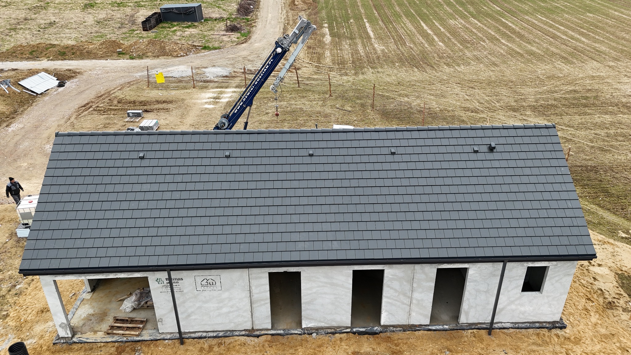 Nowoczesny dom w trakcie budowy z ciemnoszarą dachówką karpiówką, widok z góry. Na dachu dźwig budowlany. W tle pole i droga gruntowa.