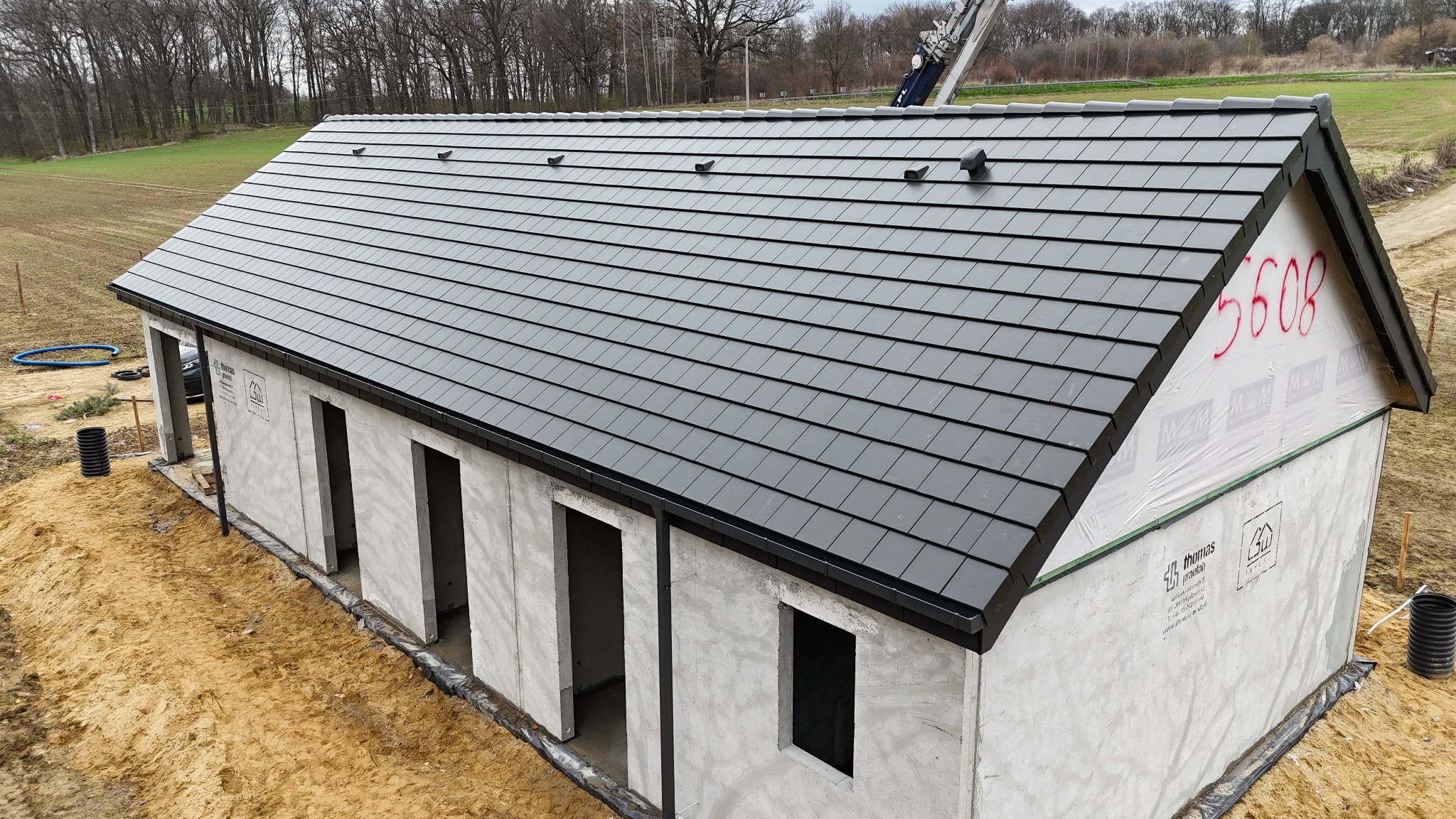 Nowoczesny, grafitowy dach z dachówki ceramicznej na surowym budynku. Widoczne detale wykończenia i numer '5608' na ścianie szczytowej. Ujęcie z góry.