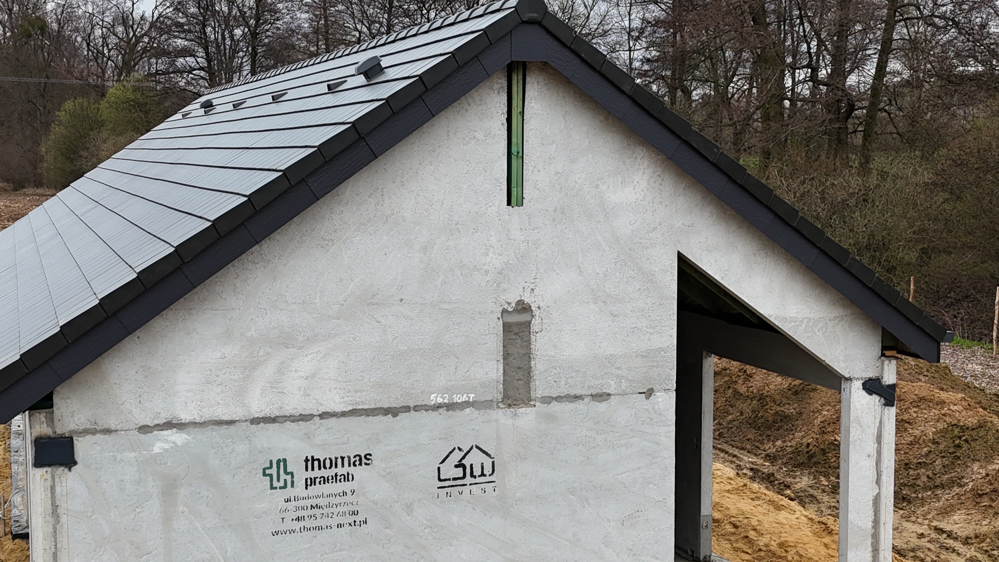 Szary dach z czarną podbitką na surowym budynku z widocznymi elementami konstrukcyjnymi i logo firmy. Widok z dołu, częściowo ukończony budynek.