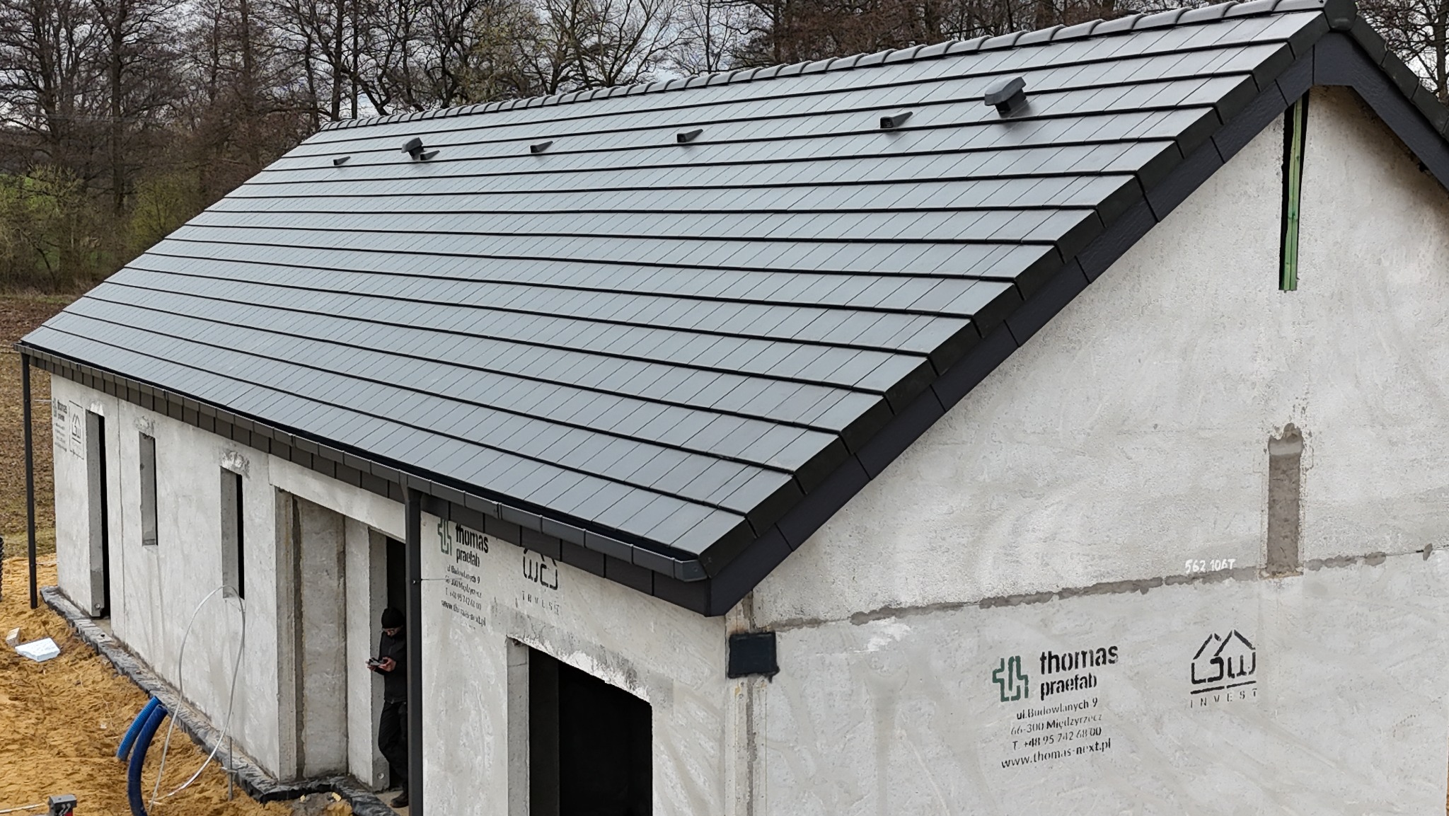 Nowy, grafitowy dach z dachówki ceramicznej na surowym budynku w trakcie budowy. Widoczne detale wykończenia i wentylacja dachu. Ujęcie z boku, w tle drzewa.