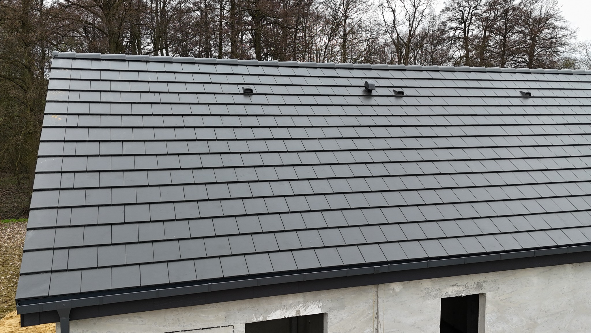 Szary dach z dachówki ceramicznej, widoczne kominki wentylacyjne. Budynek w stanie surowym zamkniętym, drzewa w tle. Ujęcie z góry, lekko z boku.