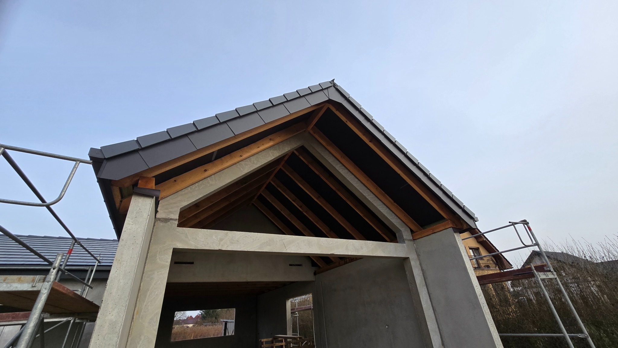 Surowa konstrukcja domu z widoczną więźbą dachową i częściowo ułożonym, grafitowym pokryciem. Widok z dołu na budynek w trakcie budowy.