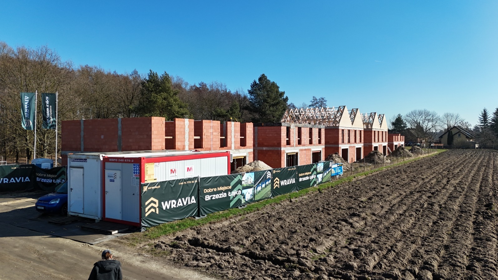 Budowa szeregówki z czerwonej cegły, widoczna więźba dachowa. Na pierwszym planie kontener budowlany i banery reklamowe osiedla Wravia Brzezia Łąka. Błękitne niebo.