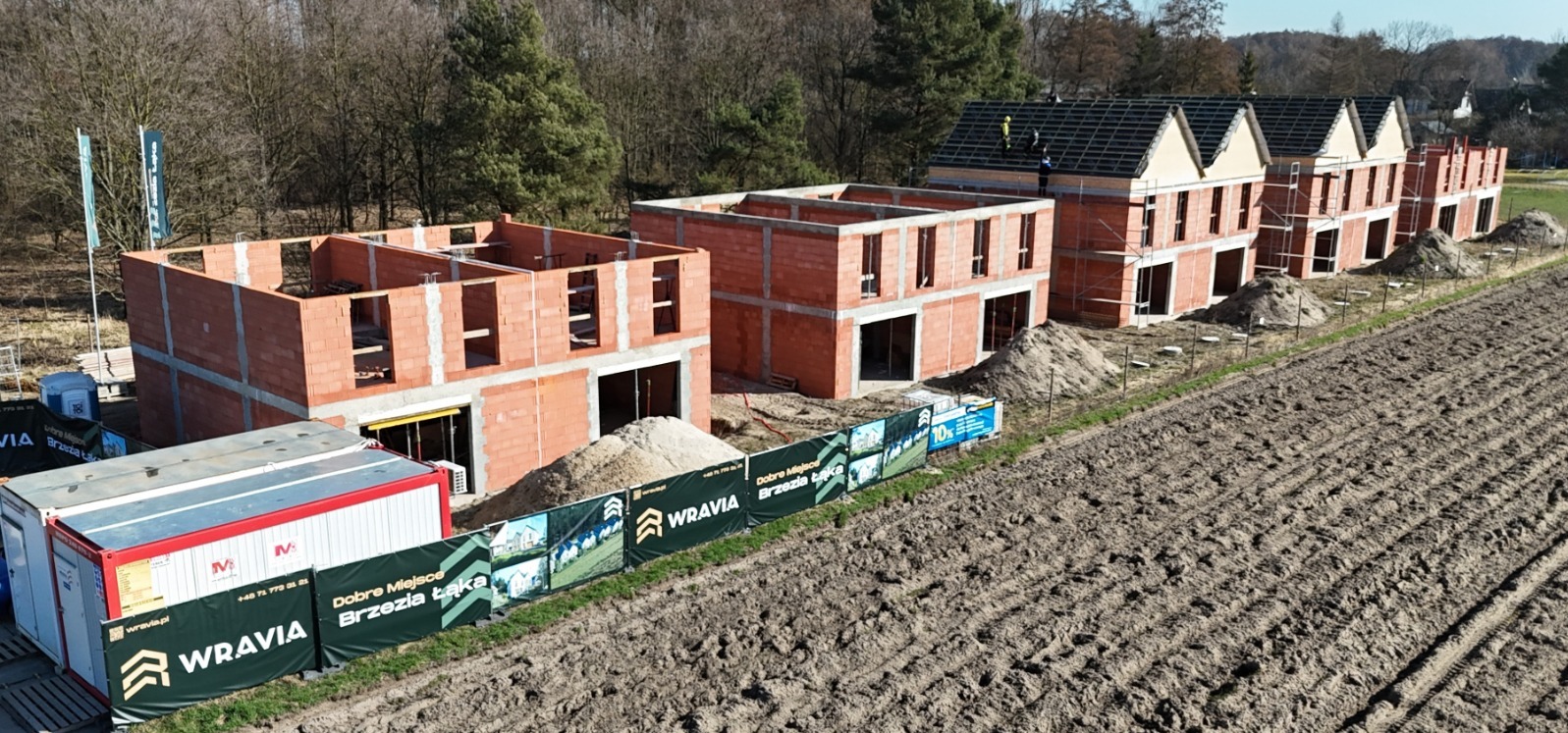 Budowa szeregu domów z czerwonej cegły w różnym stadium zaawansowania, z widoczną konstrukcją dachu na jednym z budynków. Widok z lotu ptaka na plac budowy.