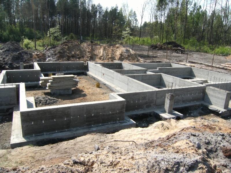 Zalane betonem fundamenty domu z widocznymi śladami po szalunkach, na placu budowy widoczny stos płyt szalunkowych i wykopana ziemia.