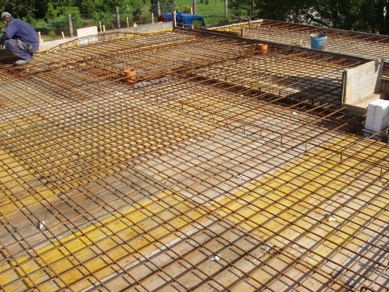 Zbrojenie fundamentu płytowego z widoczną siatką z prętów stalowych na żółtym deskowaniu, przygotowane do zalania betonem, z pracownikiem w tle.