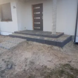 Schody wejściowe do domu wyłożone szarą kostką brukową, częściowo zasypane piaskiem, obok ułożone ażurowe płyty betonowe.