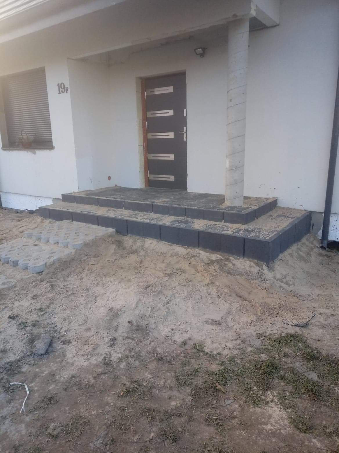 Schody wejściowe do domu wyłożone szarą kostką brukową, częściowo zasypane piaskiem, obok ułożone ażurowe płyty betonowe.