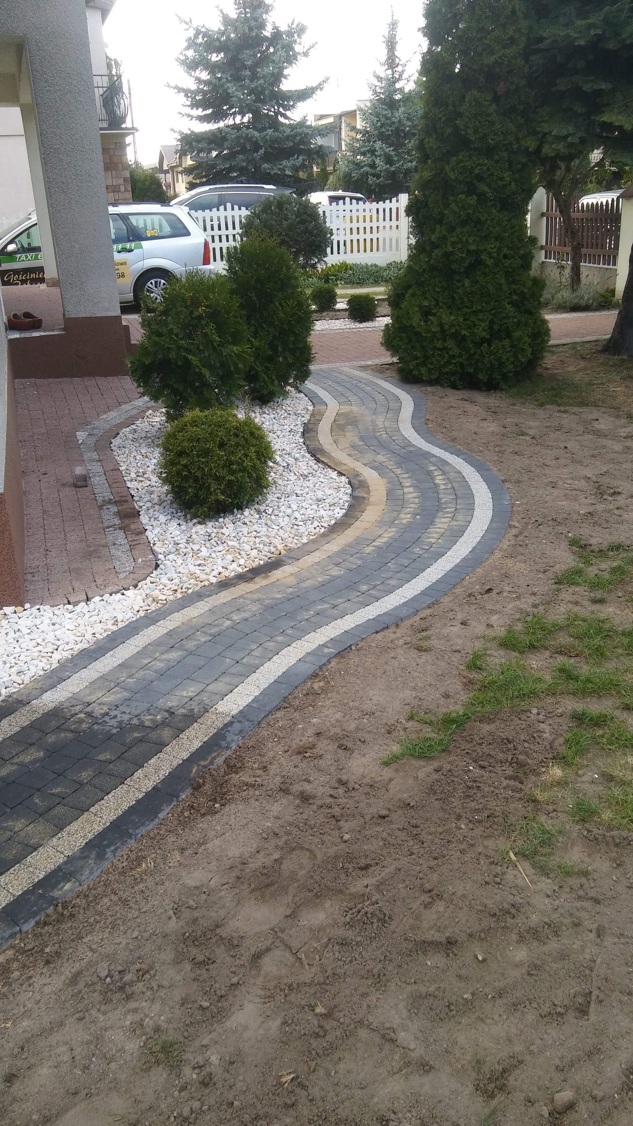 Falista ścieżka z kostki brukowej w odcieniach szarości i beżu, obrzeża z białego żwiru i zielonych krzewów, widok z góry na świeżo ułożony chodnik.