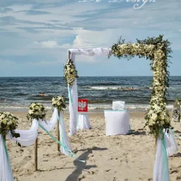 Plażowa ceremonia ślubna z białym baldachimem udekorowanym kwiatami, widok na morze i pływających ludzi w tle, flaga Polski na stoliku.