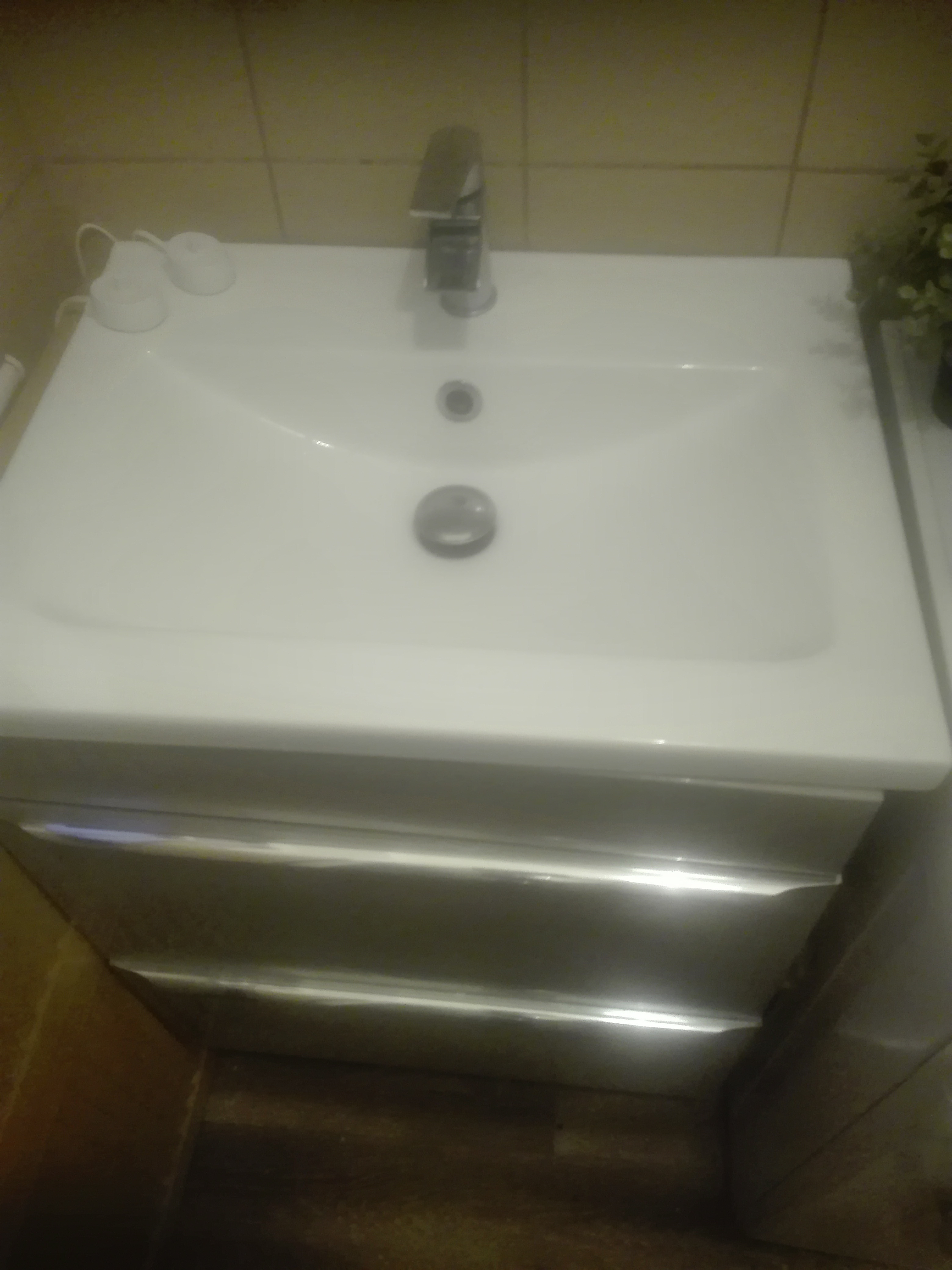 Biała umywalka z szafką i chromowaną baterią, widok z góry, w tle beżowe płytki i roślina doniczkowa.