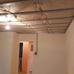 Poddasze w trakcie ocieplania: widoczne metalowe profile pod konstrukcję, wełna izolacyjna i wystające kable elektryczne, ściany w jasnym kolorze, w tle otwarte drzwi do innego pomieszczenia.