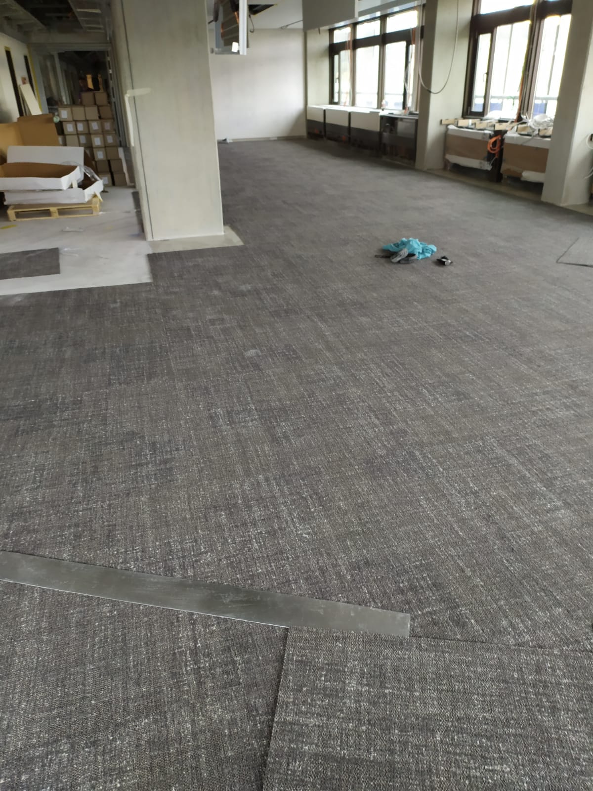 Montaż szarej wykładziny dywanowej w biurze, widoczne niedociągnięte krawędzie i metalowa listwa pomiarowa.