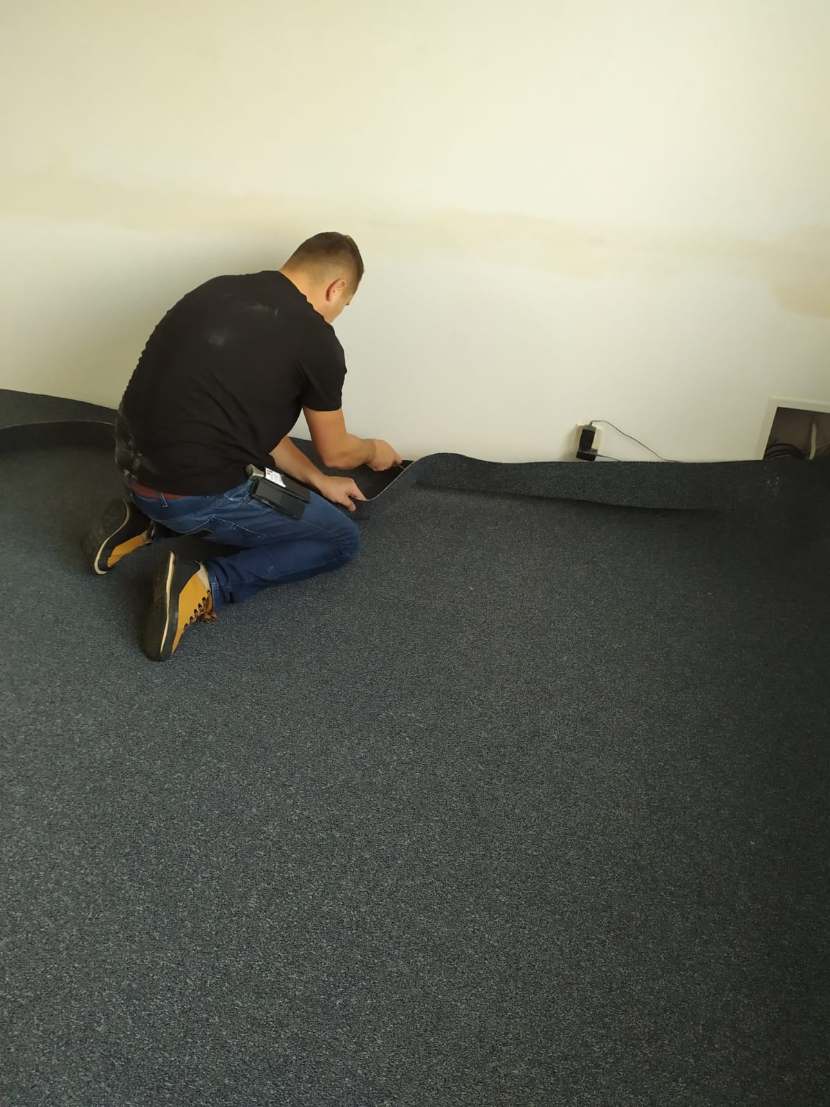 Pracownik na kolanach dopasowuje ciemnoszarą wykładzinę podłogową do ściany w pomieszczeniu ze skośnym sufitem, widoczny kabel elektryczny przy ścianie.