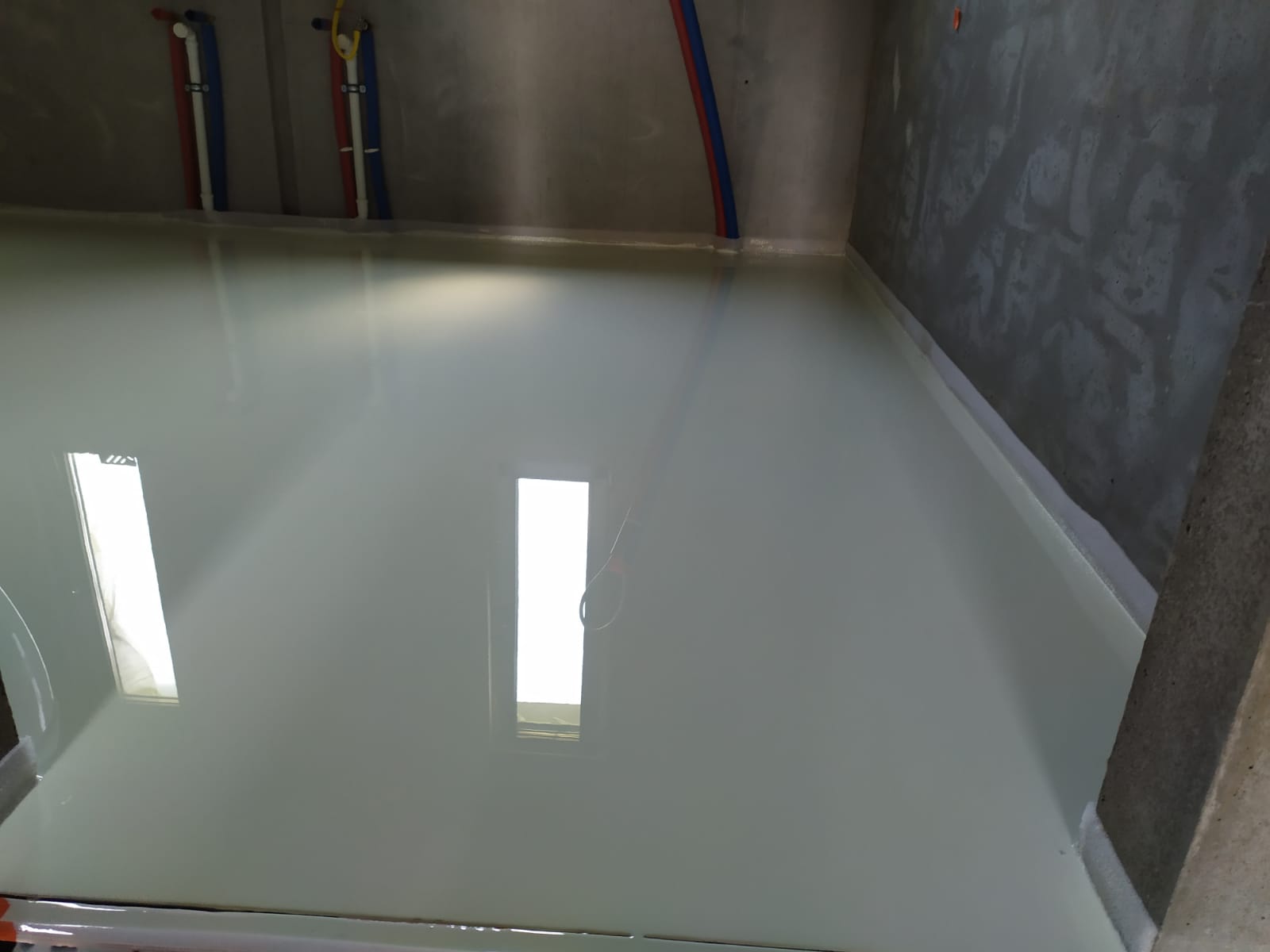 Świeżo wylana, samopoziomująca posadzka przemysłowa w szarym odcieniu, z widocznymi rurami instalacji pod sufitem i refleksami okien na powierzchni.