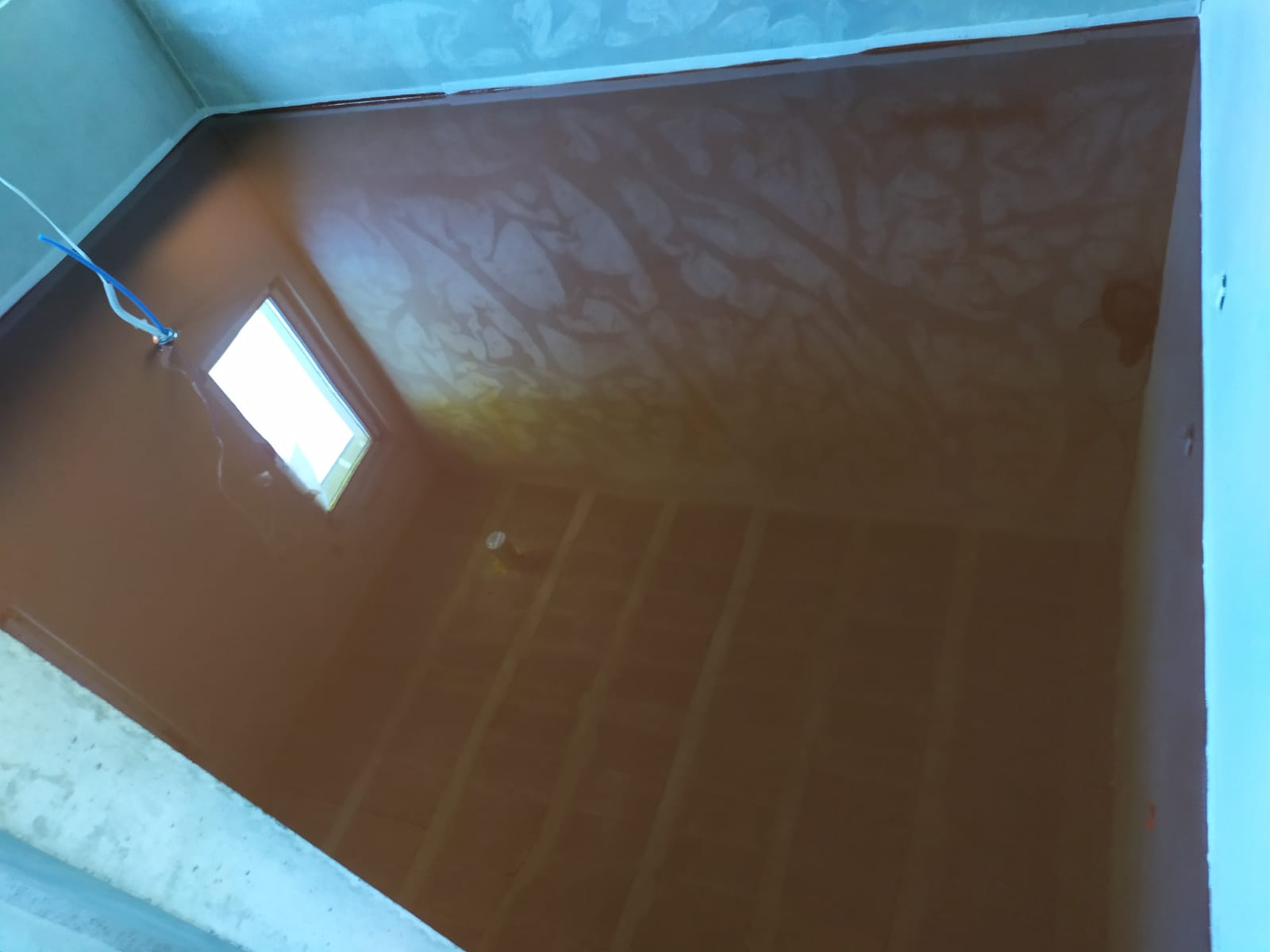 Świeżo wylana, brązowa posadzka przemysłowa w surowym wnętrzu, widoczne kable i małe okno w ścianie.