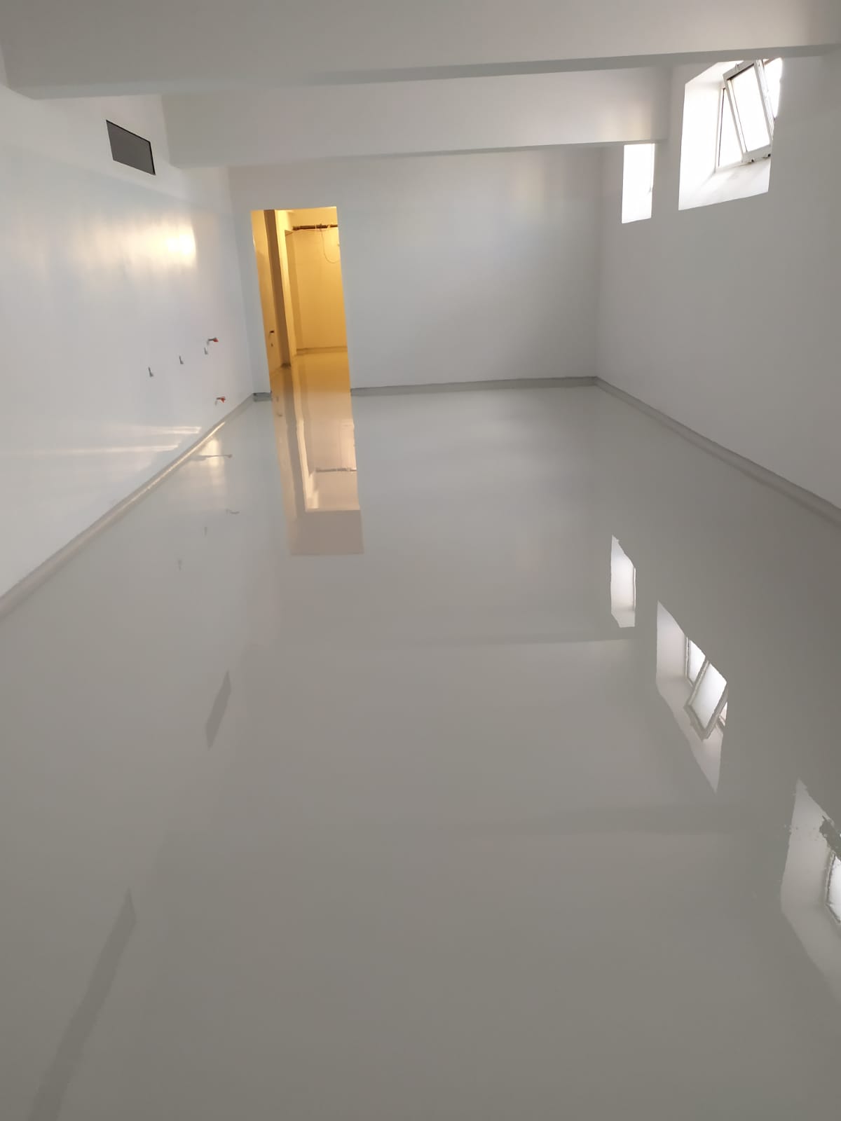 Jasnoszara, błyszcząca posadzka przemysłowa w pustym pomieszczeniu z białymi ścianami i oknami, widoczne odbicie okien w podłodze.