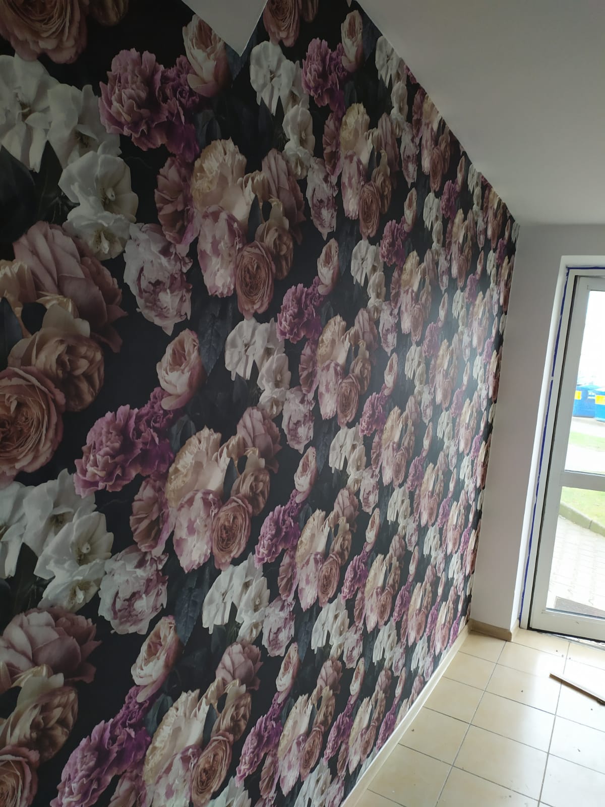 Ściana pokryta ciemną tapetą w kwiaty w odcieniach różu, fioletu i bieli, widoczna z perspektywy pod kątem, z fragmentem białej ściany i okna po prawej stronie.